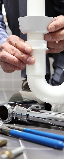 Plomberie : Installation professionnelle de siphon et évacuation. Mains d'un plombier ajustant un siphon de lavabo en PVC blanc. Tuyaux chromés et outils de serrage sur sol carrelé.